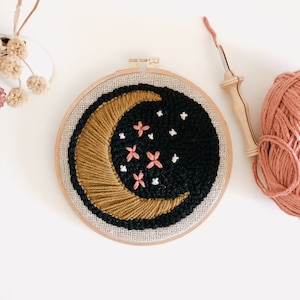 Op de afbeelding: Een gouden halve maan met roze bloemen en witte sterren geborduurd op een zwarte achtergrond. De borduurwerk is ingelijst in een natuurlijke houten ring.