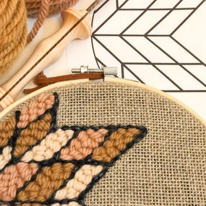 Puede incluir: Primer plano de un proyecto de bordado en forma de estrella en progreso sobre una tela de arpillera estirada en un aro de bordado de madera. La estrella est&aacute; hecha con hilo de color marr&oacute;n, beige y crema.