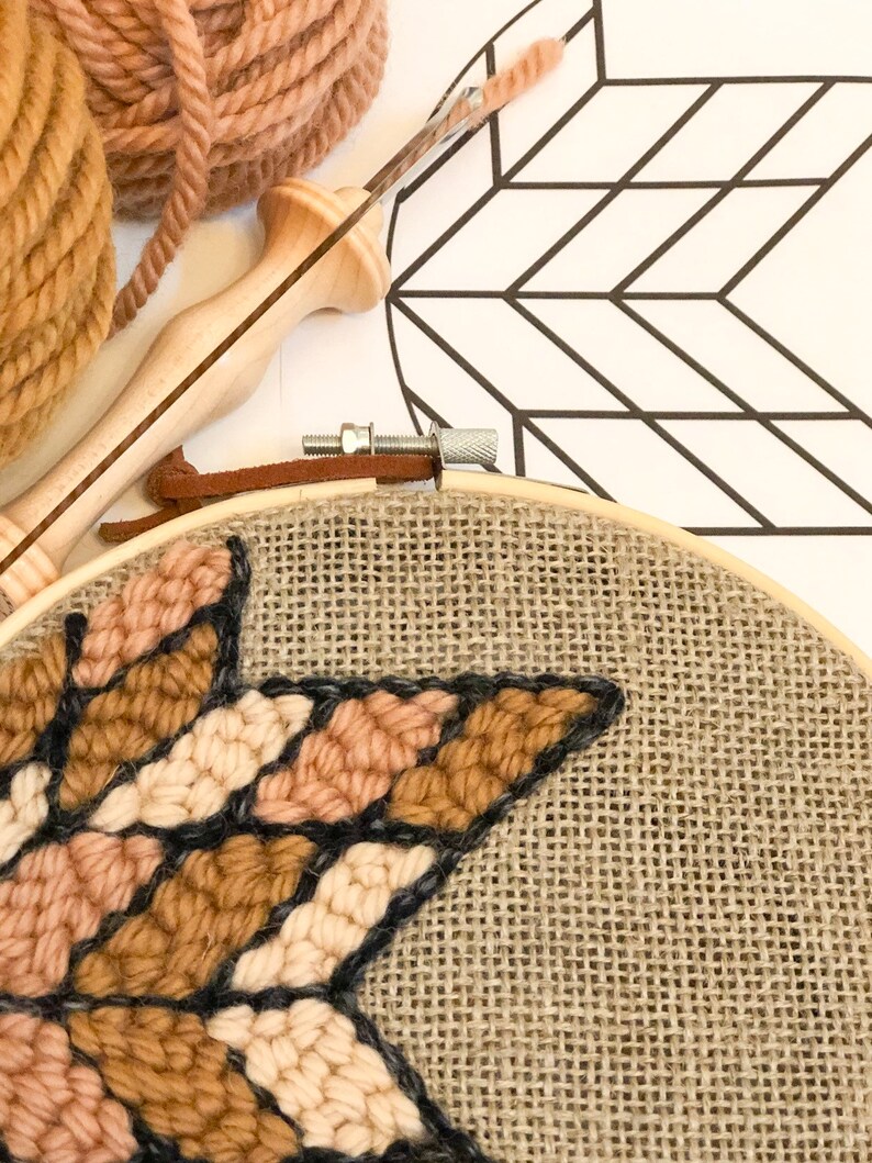 Puede incluir: Primer plano de un proyecto de bordado en forma de estrella en progreso sobre una tela de arpillera estirada en un aro de bordado de madera. La estrella est&aacute; hecha con hilo de color marr&oacute;n, beige y crema.