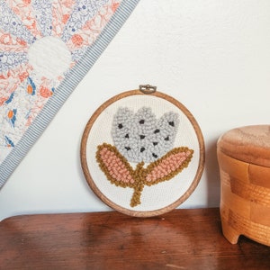 May include: A gray and white flower with pink and yellow leaves is embroidered on a white canvas in a wooden hoop.
