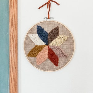May include: A circular wall hanging with a starburst design made of yarn in shades of brown, beige, pink, and blue. The yarn is woven into a beige fabric background.