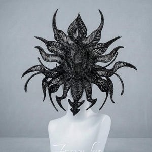 Peut inclure: Une coiffe noire composée d'une structure complexe et multicouche de fins filaments semblables à du fil de fer, disposés en un motif radial organique avec des extensions pointues et incurvées. Le texte "Fraise au Loup" est imprimé en blanc.