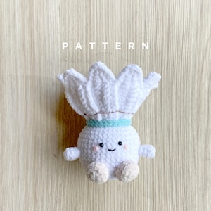 Puede incluir: Un juguete de peluche blanco tejido a mano con una cara sonriente, brazos pequeños y pies beige. El juguete tiene una parte superior en forma de corona y una banda azul claro alrededor del cuello. La palabra "PATTERN" está impresa encima del juguete.