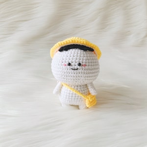 May include: White crocheted amigurumi doll with a yellow hat and a yellow bag. The doll has a black mouth and black eyes with pink cheeks.