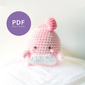 May include: A pink crocheted amigurumi toy with a white face and black eyes. The toy has a small pink pom-pom on top of its head. The toy is sitting on a white fluffy surface. PDF PATTERN.