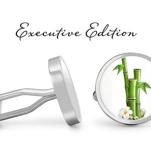 Può includere: Gemelli d'argento con sfondo bianco e un design di bambù verde con un fiore bianco. I gemelli sono etichettati "Executive Edition".