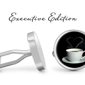 Pode incluir: Um par de botões de punho de prata com uma imagem em preto e branco de uma xícara de café com vapor em forma de coração subindo dela. Os botões de punho são etiquetados como "Executive Edition".