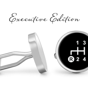 May include: A pair of silver cufflinks with a black background and white gear shift pattern. The cufflinks say "Executive Edition" on the top.