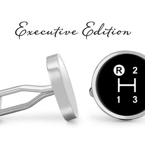 May include: A pair of silver cufflinks with a black circular design featuring a white gear shift pattern with the letters "R", "H", "1", "2", and "3". The cufflinks are labeled "Executive Edition".