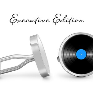 Op de afbeelding: Een paar zilveren manchetknopen met een zwart-wit vinylplaatontwerp. De plaat heeft een blauw centraal label. De manchetknopen zijn gelabeld "Executive Edition".