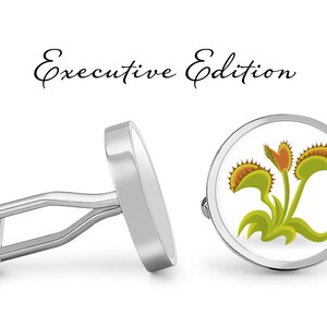 Op de afbeelding: Een paar zilveren manchetknopen met een witte achtergrond en een afbeelding van drie groene Venusvliegenvallen met oranje en gele accenten. De manchetknopen zijn gelabeld met "Executive Edition".