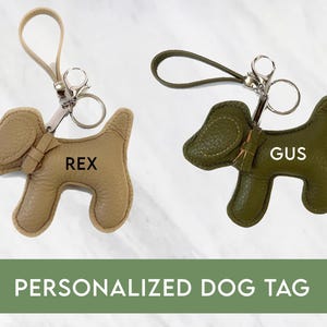 May include: Two leather dog tags in the shape of a dog, one tan and one olive green. The tan tag has the name "REX" and the green tag has the name "GUS". Both tags have a silver clasp and a loop. The text "PERSONALIZED DOG TAG" is below the tags.