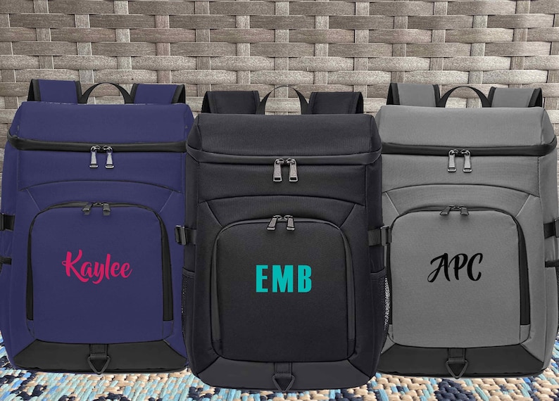 May include: Three personalized backpacks in different colors. The blue backpack has the name "Kaylee" embroidered in pink. The black backpack has the initials "EMB" embroidered in teal. The gray backpack has the initials "APC" embroidered in black.