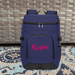 May include: A navy blue backpack with black accents and a zippered front pocket. The backpack has a pink embroidered name "Kaylee" on the front.