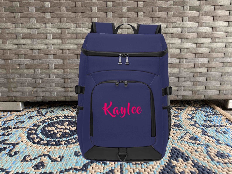 May include: A navy blue backpack with black accents and a zippered front pocket. The backpack has a pink embroidered name "Kaylee" on the front.