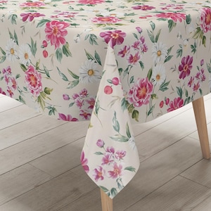 Peut inclure: Une nappe beige ornée d'un motif floral avec des pivoines roses, des marguerites blanches et des feuilles vertes. La nappe est drapée sur une table en bois, mettant en valeur le design botanique détaillé.