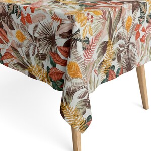 Peut inclure: Une nappe blanche à imprimé tropical avec des feuilles brunes, orange et jaunes. La nappe est drapée sur une table avec des pieds en bois.