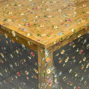 Può includere: Tovaglia di plastica trasparente con un motivo floreale con piccoli fiori rosa, gialli e blu su uno sfondo marrone. La tovaglia è drappeggiata su un tavolo di legno.