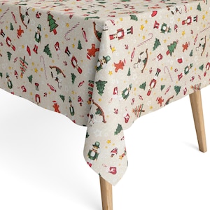 Op de afbeelding: Beige kersttafelkleed met een herhalend patroon van feestelijke elementen. Het ontwerp omvat rode notenkrakers, groene kerstbomen, peperkoekenmannen en sterren. Het tafelkleed is gedrapeerd over een houten tafel.