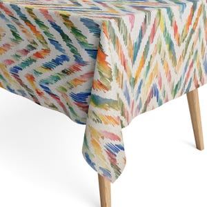 Può includere: Una tovaglia rettangolare caratterizzata da un vivace motivo a chevron multicolore con pennellate in stile acquerello nelle tonalità del blu, verde, giallo, arancione e rosa su sfondo color crema, drappeggiata su un tavolo con gambe in legno chiaro.