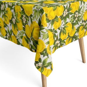 Op de afbeelding: Een wit tafelkleed met een citroenprint. Het tafelkleed heeft gele citroenen, groene bladeren en witte bloemen.