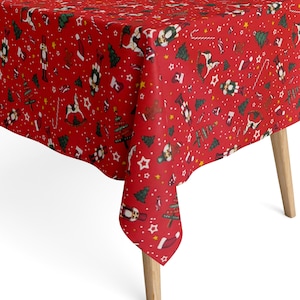 Op de afbeelding: Een rode kersttafelkleed met een feestelijk patroon. Het ontwerp bevat witte sterren, groene kerstbomen, speelgoedsoldaten en andere illustraties met een vakantiethema. Het tafelkleed is over een houten tafel gedrapeerd.