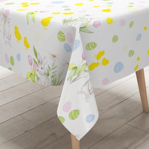 Op de afbeelding: Witte paastafelkleed met aquarel illustraties van konijnen, kuikens, eieren en bloemen. Het ontwerp bevat roze tulpen, madeliefjes en groen blad, met pastelkleurige eieren verspreid over het oppervlak. Het tafelkleed is over een houten tafel gedrapeerd.
