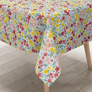 Puede incluir: Un mantel blanco con un vibrante estampado floral. El diseño presenta un estampado integral de flores rojas, amarillas y color melocotón con hojas verdes. El mantel está sobre una mesa de madera, mostrando el diseño de la tela.