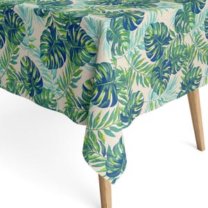 Könnte beinhalten: Ein rechteckiges Tischtuch mit einem tropischen Blättermuster in Grün und Blau auf cremefarbenem Hintergrund. Das Tischtuch ist über einem Holztisch drapiert und zeigt das lebendige Design. Die detaillierten Blätter erzeugen eine frische, natürliche Ästhetik.