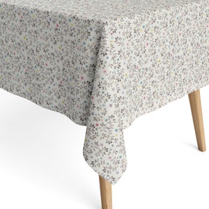 Può includere: Una tovaglia bianca con un motivo floreale con piccoli rami grigi e fiori colorati. La tovaglia è drappeggiata su un tavolo di legno con quattro gambe.