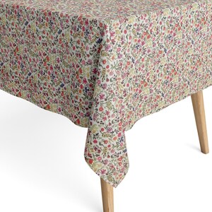 Può includere: Una tovaglia rettangolare caratterizzata da un fitto motivo floreale ripetuto con piccoli fiori nelle tonalità del rosso, rosa, giallo e blu su sfondo bianco, drappeggiata su un tavolo con gambe in legno chiaro.