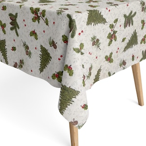 Op de afbeelding: Een witte tafelkleed met een kerstthema patroon met groene dennenbomen, rode bessen en dennenappels. De doek is over een houten tafel gedrapeerd, wat een feestelijke setting creëert.