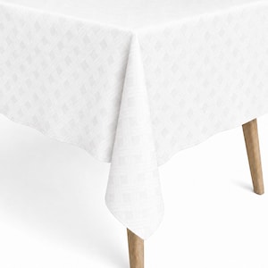 Op de afbeelding: Een wit tafelkleed met een subtiel, geweven mandpatroon, gedrapeerd over een tafel met lichtbruine houten poten. De stof heeft een herhalende geometrische textuur die een zacht, monochroom visueel effect over het hele oppervlak creëert.