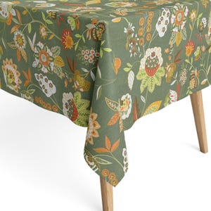 Könnte beinhalten: Ein grünes Tischtuch mit floralem Muster. Das Design zeigt große Blüten in Weiß, Orange und Gelb auf grünem Hintergrund. Das Tischtuch ist über einem Holztisch drapiert.