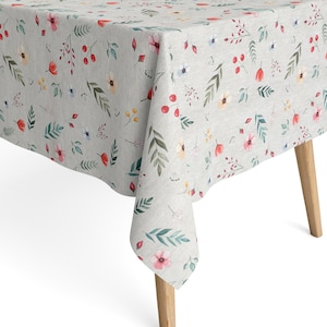 Può includere: Una tovaglia grigio chiaro con un motivo floreale. Il design presenta fiori in stile acquerello nei toni del rosa, rosso, giallo e blu, insieme a foglie e bacche verdi. La tovaglia è drappeggiata su un tavolo di legno.