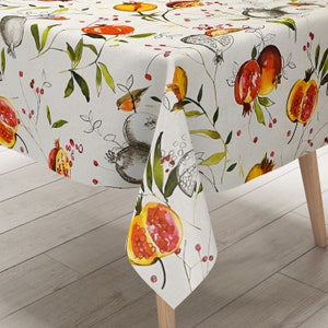 Peut inclure: Une nappe blanche avec un motif de grenade et de feuilles. La nappe est en mélange de lin et a un léger brillant. La nappe est présentée drapé sur une table avec des pieds en bois.