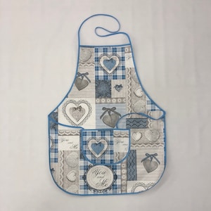 Op de afbeelding: Een blauwe en grijze patchwork schort met een grote zak en de tekst "You and Me" in een cirkel. Het schort heeft een blauwe rand en een hartjespatroon.
