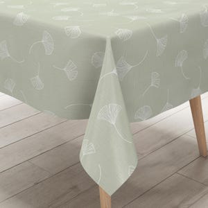 Può includere: Una tovaglia verde salvia con un motivo ripetuto di foglie di ginkgo bianche. La tovaglia è drappeggiata su un tavolo di legno, mostrando la trama e il design del tessuto. Il motivo a foglie di ginkgo aggiunge un tocco di eleganza ispirata alla natura alla tavola.