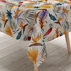 Op de afbeelding: Een beige tafelkleed met een tropische print met grote oranje, gele en rode bladeren en bloemen. Het tafelkleed is over een houten tafel gelegd.