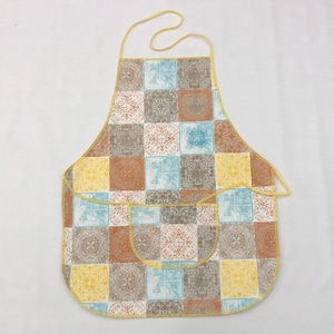 Op de afbeelding: Een gele en bruine gepatroneerde schort met twee zakken. Het schort heeft een geruit ontwerp met blauwe, bruine en gele vierkanten.