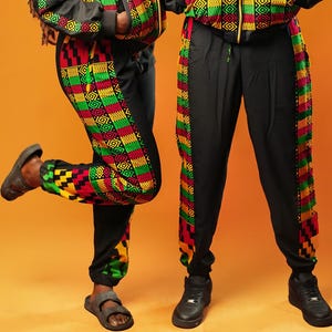 Woman and man standing against an orange background wearing black tracksuit trousers with colourful Kente African print panels down the sides. The trousers have a relaxed fit with elastic cuffs at the ankles and a drawstring waist. They both pair them with matching black jackets featuring Kente trim.