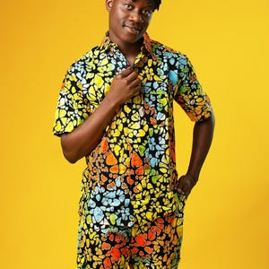A man stands in front of a yellow background wearing Funky Men’s Cargo Shorts made from multicoloured African print fabric with a butterfly pattern in orange, yellow, blue, and black. The shorts have a relaxed fit and side pockets, paired with a matching short-sleeve shirt. This is a bold Summer Hawaiian Shorts look, ideal for African Print Men’s Clothing, a Hippie Festival Set, or a Carnival Outfit from Ashanti Empress.