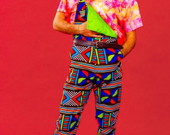 May include: A person wearing a pink and white tie-dye shirt with a green bandana, a brown belt, and black and blue patterned pants. The person is wearing black sunglasses and has long curly hair.