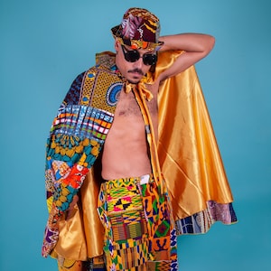 Op de afbeelding: Een persoon draagt een kleurrijke, gepatroneerde cape en shorts met een gouden hoed en zonnebril. De cape is geel en heeft een gepatroneerde zoom. De shorts zijn gemaakt van een kleurrijke stof met een geometrisch patroon. De hoed is een brede hoed met een patroon.