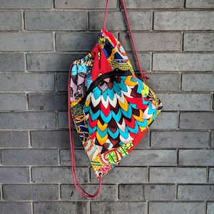 Peut inclure: Un sac à cordon coloré en tissu patchwork avec un motif rouge, bleu, jaune et blanc. Le sac est suspendu à un mur de briques.