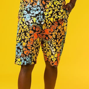 A man stands in front of a yellow background wearing Funky Men’s Cargo Shorts made from multicoloured African print fabric with a butterfly pattern in orange, yellow, blue, and black. The shorts have a relaxed fit and side pockets, paired with a matching short-sleeve shirt. This is a bold Summer Hawaiian Shorts look, ideal for African Print Men’s Clothing, a Hippie Festival Set, or a Carnival Outfit from Ashanti Empress.