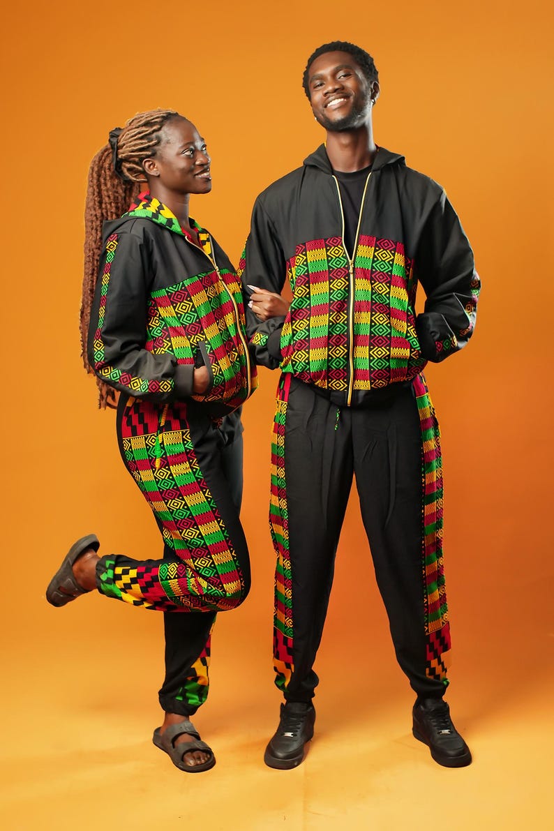 Woman and man standing against an orange background wearing black tracksuit trousers with colourful Kente African print panels down the sides. The trousers have a relaxed fit with elastic cuffs at the ankles and a drawstring waist. They both pair them with matching black jackets featuring Kente trim.