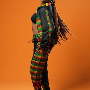 Woman standing against an orange background wearing black tracksuit trousers with colourful Kente African print panels down the sides. The trousers have a relaxed fit with elastic cuffs at the ankles and a drawstring waist. She pairs them with a matching black jacket featuring Kente trim and a black crop top.