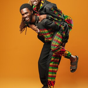 Woman and man standing against an orange background wearing black tracksuit trousers with colourful Kente African print panels down the sides. The trousers have a relaxed fit with elastic cuffs at the ankles and a drawstring waist. They both pair them with matching black jackets featuring Kente trim.