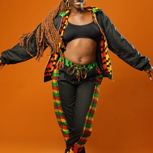Woman standing against an orange background wearing black tracksuit trousers with colourful Kente African print panels down the sides. The trousers have a relaxed fit with elastic cuffs at the ankles and a drawstring waist. She pairs them with a matching black jacket featuring Kente trim and a black crop top.